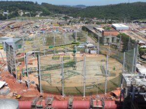 An industrial construction site with large cylindrical structures, cranes, and scaffolding, surrounded by netting and equipment, set in a hilly area with trees and buildings in the background.