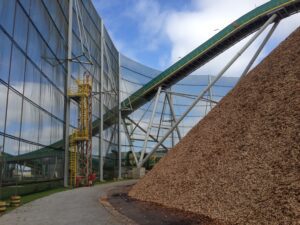 Large pile of wood chips next to an industrial conveyor belt and metal structure, with tall safety netting and a partly cloudy sky overhead.